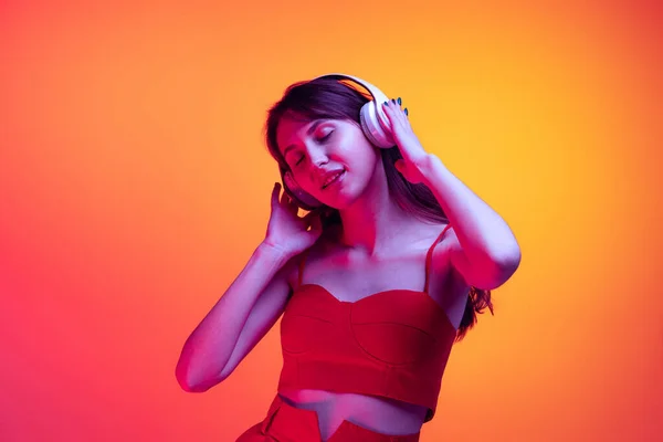 Портрет молодої дівчини в навушниках, що слухає музику ізольовано на червоно-жовтому фоні в неоновому світлі. Поняття емоцій, вираження обличчя, мистецтво, краса — стокове фото
