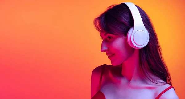 Ritratto di giovane ragazza in cuffia che ascolta musica isolata su sfondo rosso-giallo in luce al neon. Concetto di emozioni, espressione facciale, arte, bellezza — Foto Stock