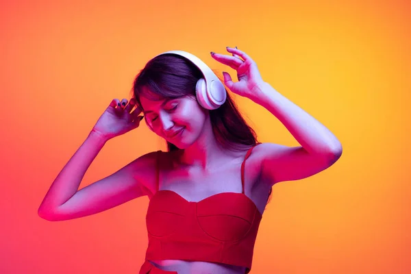 Портрет молодої дівчини в навушниках, що слухає музику ізольовано на червоно-жовтому фоні в неоновому світлі. Поняття емоцій, вираження обличчя, мистецтво, краса — стокове фото