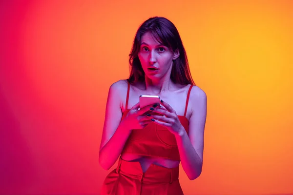 Portret van jong emotioneel meisje, student met behulp van telefoon geïsoleerd over oranje studio achtergrond in neon licht. Concept van emoties, gezichtsuitdrukking, kunst, schoonheid — Stockfoto