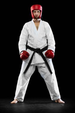 Portrait of male taekwondo fighter in white dobok, helmet and gloves posing isolated over dark background. Concept of sport, workout, competition, ad
