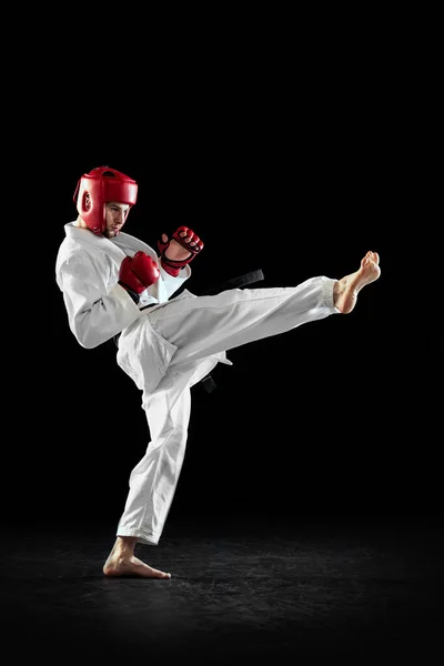 Male taekwondo fighter in white dobok, helmet and gloves training isolated over dark background. Concept of sport, workout, competition, ad — Stock Photo, Image