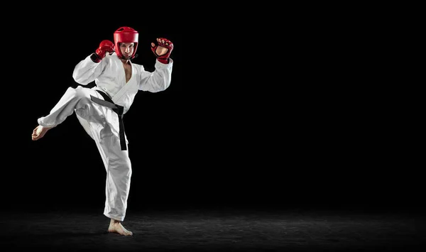 Male taekwondo fighter in white dobok, helmet and gloves training isolated over dark background. Concept of sport, workout, competition, ad — Stock Fotó