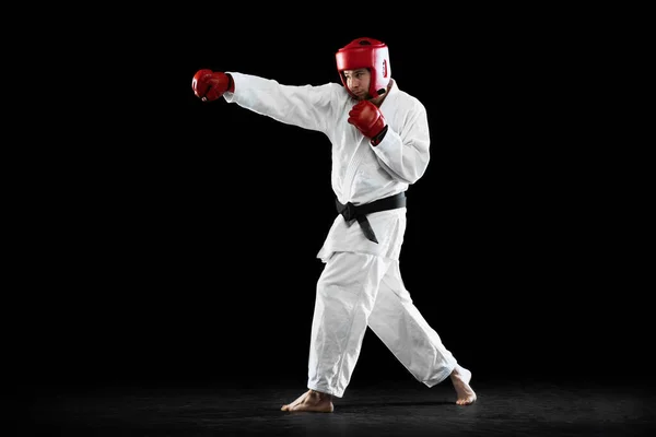 Male taekwondo fighter in white dobok, helmet and gloves training isolated over dark background. Concept of sport, workout, competition, ad — Photo