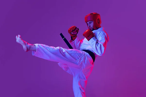 Студійний знімок чоловічого тхеквондо-бійця в спортивній формі в дії ізольовано на фіолетовому фоні в неоновому світлі. Концепція спорту, тренування, змагання, реклама — стокове фото