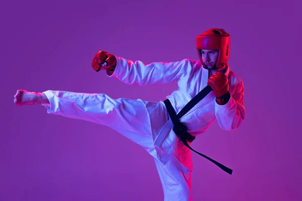 Studio shot of male taekwondo fighter in sports uniform in action isolated over purple background in neon light. Concept of sport, workout, competition, ad — 스톡 사진