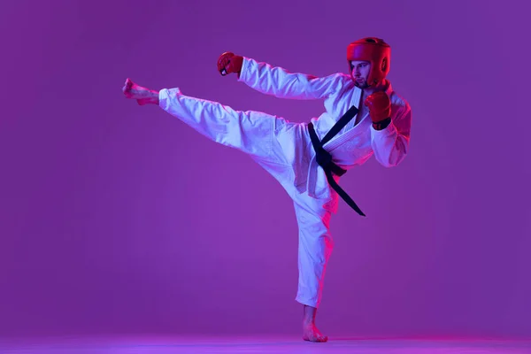 Estúdio tiro de lutador de taekwondo masculino em uniforme esportivo em ação isolado sobre fundo roxo em luz de néon. Conceito de esporte, treino, competição, anúncio — Fotografia de Stock