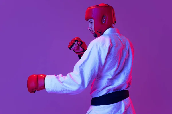 Студійний знімок чоловічого тхеквондо-бійця в спортивній формі в дії ізольовано на фіолетовому фоні в неоновому світлі. Концепція спорту, тренування, змагання, реклама — стокове фото