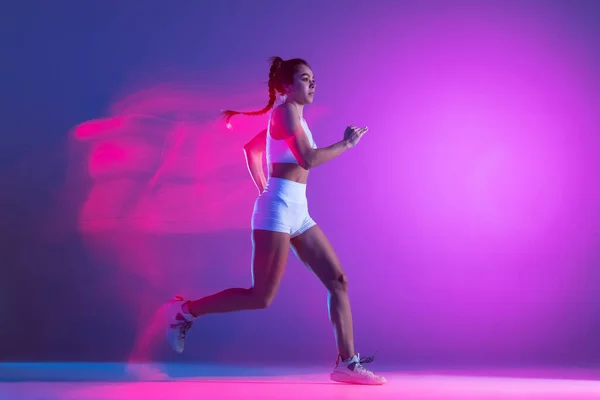 Atleta feminina profissional, treinamento de corredor isolado no fundo do estúdio azul em luz de néon rosa mista. Estilo de vida saudável, esporte, movimento e conceito de ação. — Fotografia de Stock