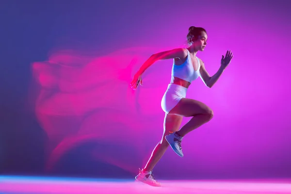 Professional female athlete, runner training isolated on blue studio background in mixed pink neon light. Healthy lifestyle, sport, motion and action concept. — Stok Foto