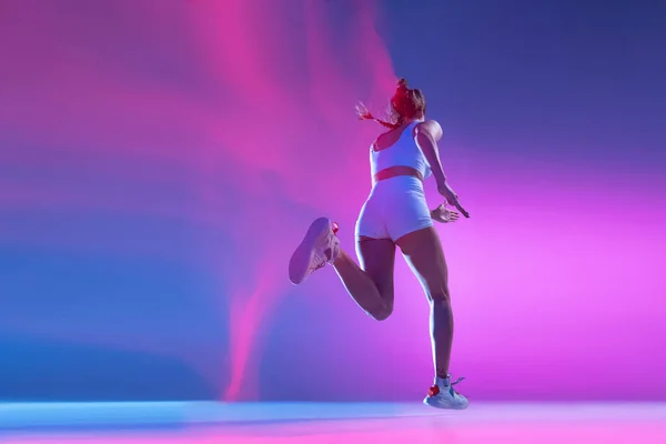 Back view. Professional female athlete running away isolated on blue studio background in mixed pink neon light. Healthy lifestyle, sport, motion and action concept. — Stock fotografie