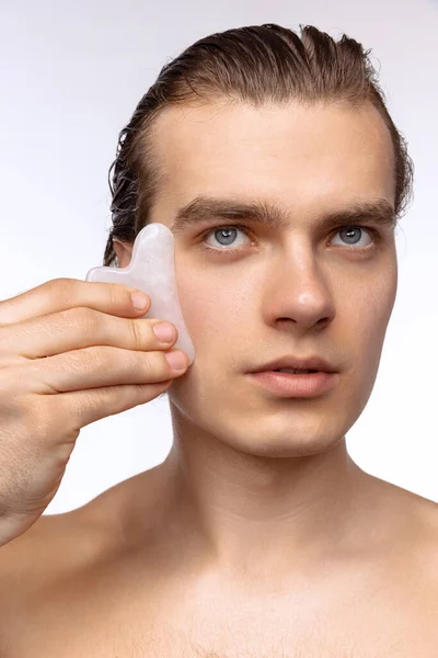 完璧な滑らかな肌を持つ若い魅力的な男は、白いスタジオの背景に隔離された顔マッサージを適用します。化粧品、健康管理、スキンケア、美しさ — ストック写真