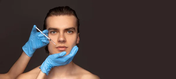 Close-up face of young man and female hands in blue gloves with syringe isolated on dark background. Cosmetic, filling surgery procedures. Anti-aging concept. — Stockfoto