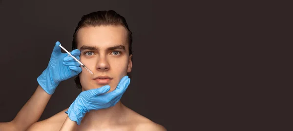 Cara de perto de homem jovem e mãos femininas em luvas azuis com seringa isolada em fundo escuro. Cosméticos, procedimentos de cirurgia de enchimento. Conceito anti-envelhecimento. — Fotografia de Stock