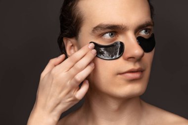 Young adorable man with well-kept skin using patches under eyes isolated over grey background. Fashion, cosmetics, health care, skin care, beauty