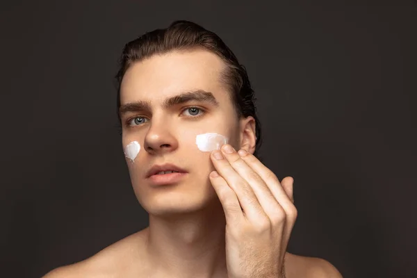 Facial skin care for man. Young happy man with perfect well-kept skin using face cream isolated over grey background. Cosmetics, health care, skin care, beauty — ストック写真