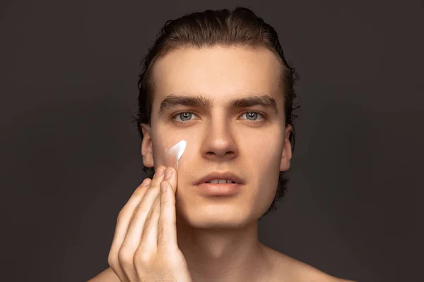 Facial skin care for man. Young happy man with perfect well-kept skin using face cream isolated over grey background. Cosmetics, health care, skin care, beauty — ストック写真