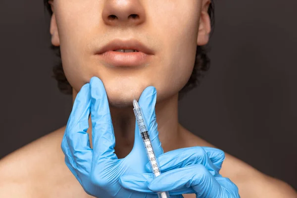 Close-up face of young man and female hands in blue gloves with syringe isolated on dark background. Cosmetic, filling surgery procedures. Anti-aging concept. — Fotografia de Stock