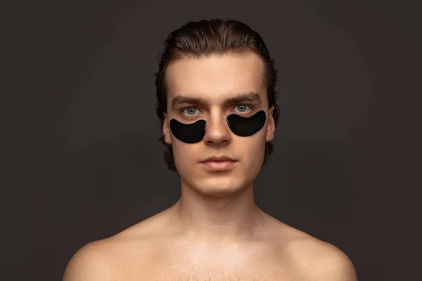 Homem jovem adorável com pele bem cuidada usando manchas sob os olhos isolados sobre fundo cinza. Moda, cosméticos, cuidados de saúde, cuidados com a pele, beleza — Fotografia de Stock