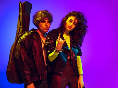 Siyah deri kıyafetler giyen, el kol hareketi yapan, bağıran ve mavi sarı arka planda neon ışıkta poz veren iki genç rock 'n' roll müzisyeni.