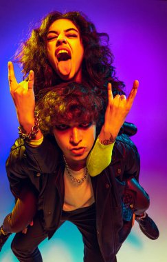 Rock music fans. Excited young man and girl in black leather outfits gesturing, shouting and dancing on blue- yellow background in neon light