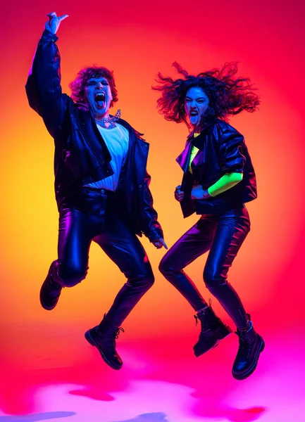 Excited young man and girl in black leather outfits having fun, gesturing, shouting and shaking head over yellow-red background in neon light — Stock fotografie