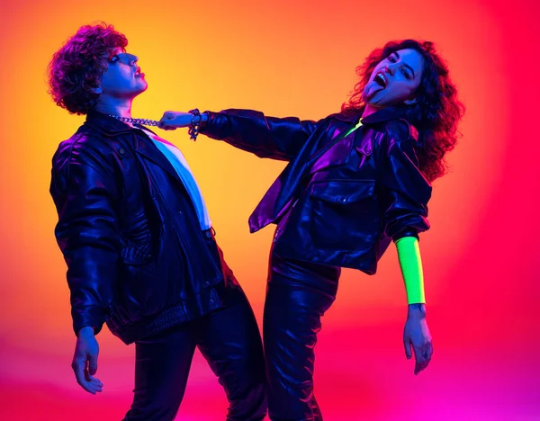 Excited young man and girl in black leather outfits having fun, gesturing, shouting and shaking head over yellow-red background in neon light — Foto Stock