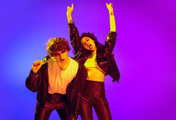 Rock music fans. Excited young man and girl in black leather outfits gesturing, shouting and dancing on blue- yellow background in neon light — Stock fotografie