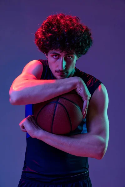Retrato de un joven jugador de baloncesto muscular mirando a la cámara aislada sobre fondo púrpura en neón. Concepto de deporte, arte, creación, competición. — Foto de Stock