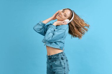 Portrait of young excited girl listening to music in headphones isolated on blue background. Concept of beauty, music, fashion, youth, spring collection