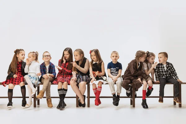 Shares secrets. Ten school and preschool age children, girls and boys sitting on banch isolated on grey background. Beauty, kids fashion, education, happy childhood concept. — Stockfoto