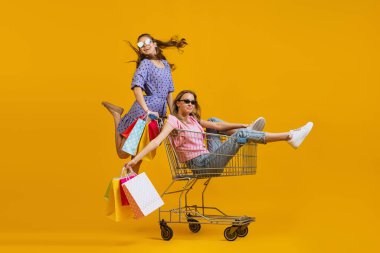 Portrait of cool young excited girls go shopping with shopping cart isolated on bright yellow background. Concept of sales, black friday, discount, emotions