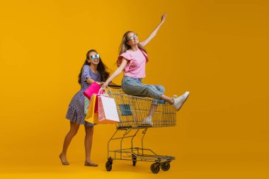 Havalı, heyecanlı genç kızların dinamik portresi alışverişe parlak sarı arka planda izole edilmiş alışveriş arabalarıyla gider. Satış kavramı, Kara Cuma, indirim, duygular.