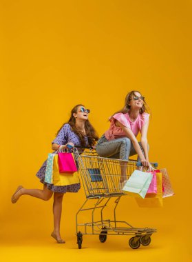Havalı, heyecanlı genç kızların dinamik portresi alışverişe parlak sarı arka planda izole edilmiş alışveriş arabalarıyla gider. Satış kavramı, Kara Cuma, indirim, duygular.