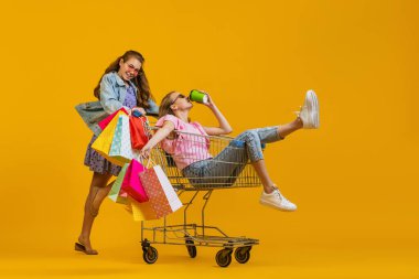 Güzel, genç, afallamış kızlar alışverişe parlak sarı arka planda izole edilmiş alışveriş arabalarıyla giderler. Satış kavramı, Kara Cuma, indirim, duygular.