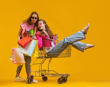 Havalı, heyecanlı genç kızların dinamik portresi alışverişe parlak sarı arka planda izole edilmiş alışveriş arabalarıyla gider. Satış kavramı, Kara Cuma, indirim, duygular.