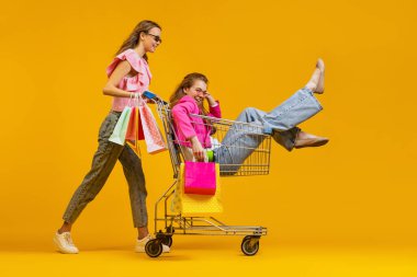 Havalı, heyecanlı genç kızların dinamik portresi alışverişe parlak sarı arka planda izole edilmiş alışveriş arabalarıyla gider. Satış kavramı, Kara Cuma, indirim, duygular.