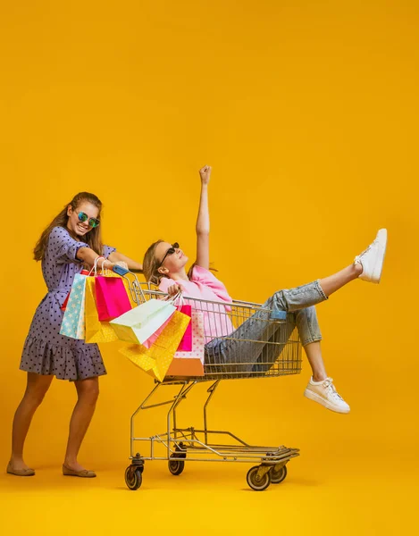 Dynamic portrait of cool young excited girls go shopping with shopping cart isolated on bright yellow background. Concept of sales, black friday, discount, emotions — Stockfoto