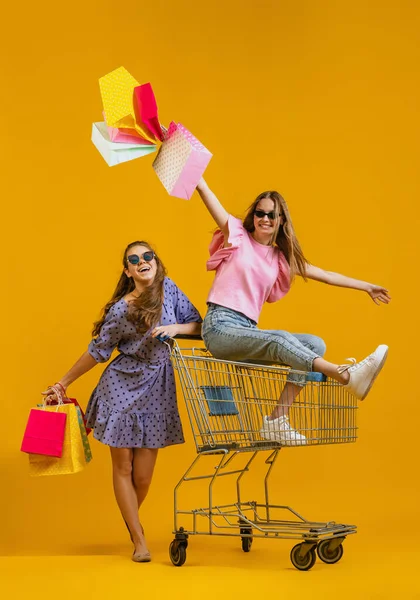 Dynamisches Porträt cooler junger aufgeregter Mädchen, die mit Einkaufswagen auf leuchtend gelbem Hintergrund einkaufen gehen. Verkaufskonzept, Schwarzer Freitag, Rabatt, Emotionen — Stockfoto