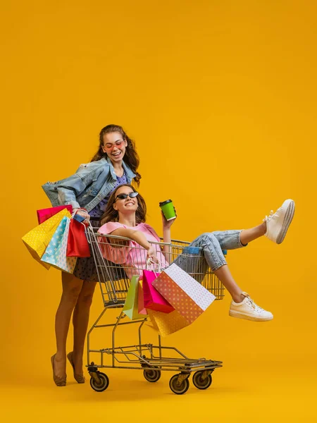 Güzel, genç, afallamış kızlar alışverişe parlak sarı arka planda izole edilmiş alışveriş arabalarıyla giderler. Satış kavramı, Kara Cuma, indirim, duygular.