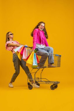 Komik alışveriş zamanı. Havalı genç mutlu kızlar alışverişe parlak sarı arka planda izole edilmiş alışveriş arabalarıyla giderler. Satış kavramı, Kara Cuma, indirim, duygular.