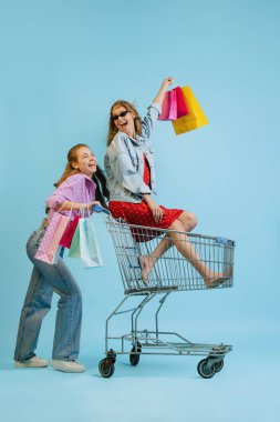 Alışveriş çantalı mutlu kızların stüdyo fotoğrafları mavi arka planda izole edilmiş bir arabaya biniyorlar. Satış kavramı, Kara Cuma, indirim, duygular, gençlik ve moda