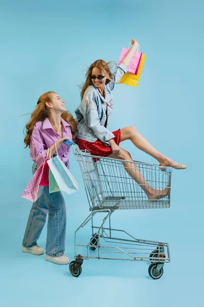 Alışveriş çantalı mutlu kızların stüdyo fotoğrafları mavi arka planda izole edilmiş bir arabaya biniyorlar. Satış kavramı, Kara Cuma, indirim, duygular, gençlik ve moda