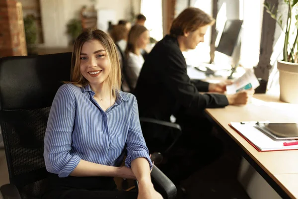Atractiva joven empresaria confiada sentada en el escritorio de la oficina con un grupo de colegas en segundo plano. Concepto de éxito, negocio, carrera, logros — Foto de Stock