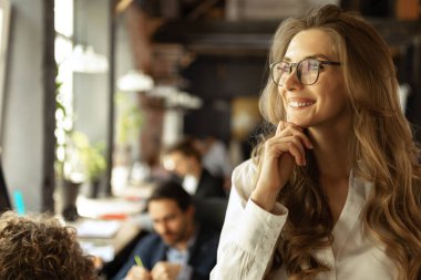 Beautiful young confident businesswoman sitting at office desk with group of colleagues in background. Concept of success, business, career, achievements