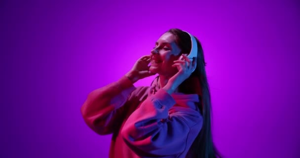 Šťastně se usmívající mladá dívka poslouchající hudbu ve sluchátkách a tančící na purpurovém pozadí v neonovém světle, filtr. 4K. Emoce, reklamy, kultura, koncepce mládeže — Stock video