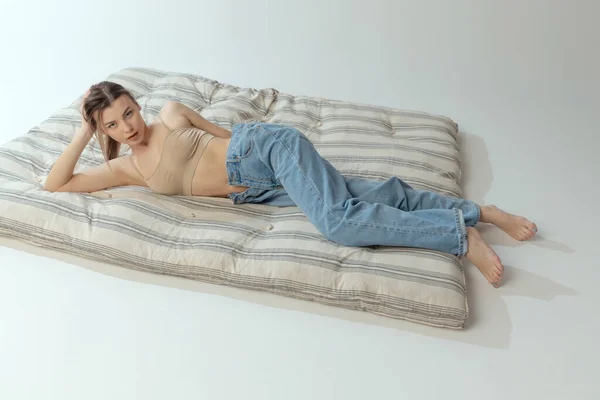 Портрет молодой привлекательной девушки, лежащей на матрасе, позирующей в бежевом нижнем белье и джинсах на фоне серой студии — стоковое фото