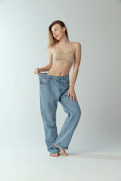 Retrato completo de mulher magra jovem vestindo jeans grandes, posando isolada em fundo de estúdio cinza. Forma perfeita do corpo. — Fotografia de Stock