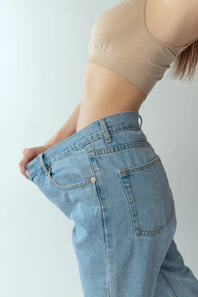 Обрізане зображення стрункої жінки в гігантських джинсах ізольовано на сірому студійному фоні. Втрата ваги, концепція дієти — стокове фото
