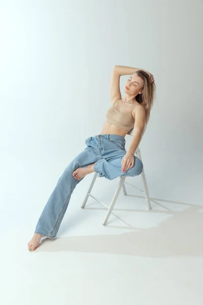 Porträtt av anbud ung flicka sitter på stol, poserar i beige underkläder och stora jeans isolerad över grå studio bakgrund. Självkärlek — Stockfoto